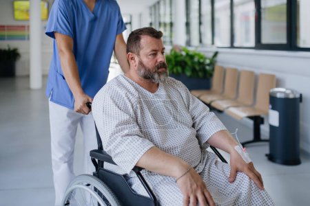 Foto de Enfermera masculina empujando a un paciente en silla de ruedas a lo largo de un pasillo del hospital, esperando un examen médico. El paciente con sobrepeso se siente ansioso y tiene problemas de salud. - Imagen libre de derechos