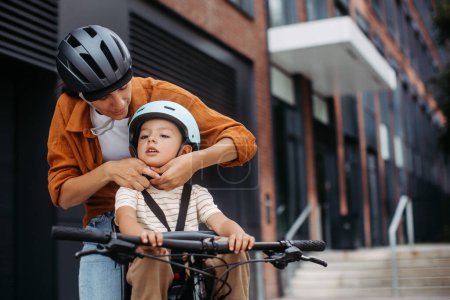 Téléchargez les photos : Mère attachant les fils casque de vélo sur la tête, le soignant sur le porte-vélo ou le siège de l'enfant. Maman faire la navette avec un jeune enfant à travers la ville sur un vélo. - en image libre de droit