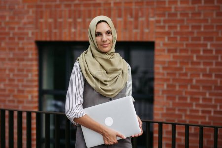 Téléchargez les photos : Portrait de belle femme en hijab debout sur la rue de la ville. Femme d'affaires musulmane avec ordinateur portable dans les mains. Iran, Afganistan enseignante devant le bâtiment de l'école. - en image libre de droit