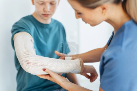 Téléchargez les photos : Médecin vérifiant le plâtre orthopédique, attelle sur un adolescent le bras cassé. Adolescent guérit une fracture après un accident. - en image libre de droit