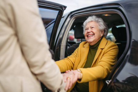 Die Seniorin steigt aus dem Auto, eine Betreuerin hilft ihr, hält ihre Hände. Ältere Frau hat Problem mit dem Aufstehen vom Rücksitz.