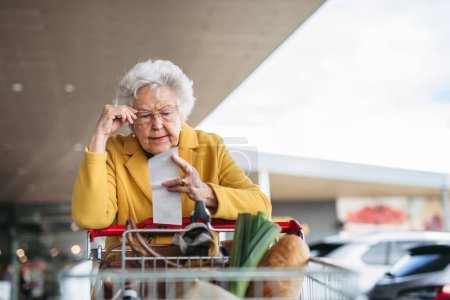 Téléchargez les photos : Une femme âgée vérifiant son reçu après un achat, regardant le montant de l'argent dépensé, s'assurant que toutes les charges sont correctes. - en image libre de droit