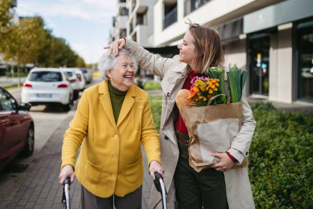 Photo for Mature granddaughter carrying grandmothers shopping bag. Senior woman and caregiver going to home with goceries from the supermarket, during cold autumn day. - Royalty Free Image
