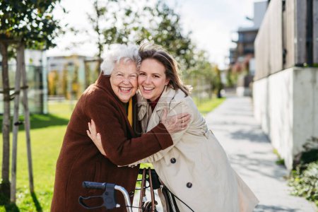 Photo for Grandmother and mature granddaughter on a walk in the city park, during a windy autumn day. Caregiver and senior lady enjoying the fall weather, hugging each other. - Royalty Free Image