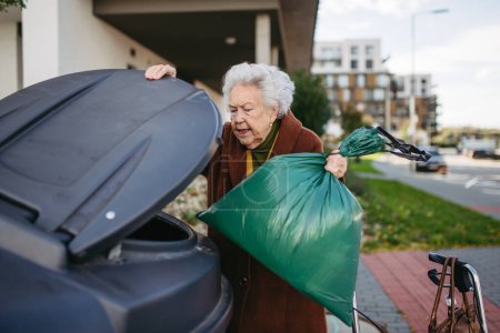 Photo for Elderly woman disposing of trash into the garbage can of waste container in front of her apartment complex. - Royalty Free Image