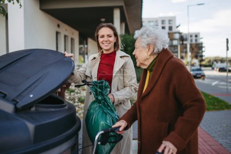 Téléchargez les photos : Femme aidant voisin âgé jeter les ordures dans la poubelle, conteneur de déchets en face de leur complexe d'appartements. - en image libre de droit