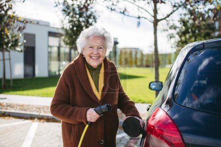 Photo for Close up of beautiful senior woman plugging charger in her electric car. Progressive elderly woman charging her electric vehicle on the street. - Royalty Free Image