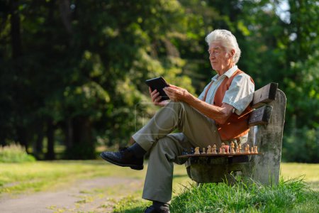 Téléchargez les photos : Homme âgé assis à l'extérieur et faisant du shopping en ligne sur une tablette numérique. Le risque d'escroqueries en ligne ciblant les personnes âgées. - en image libre de droit