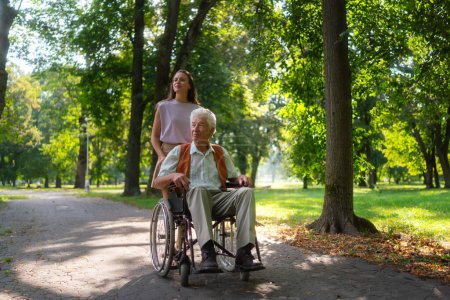 Photo for Granddaughter pushing the grandfather in wheelchair in city park. A young caregiver spending quality time with lonely senior client in the city park. - Royalty Free Image