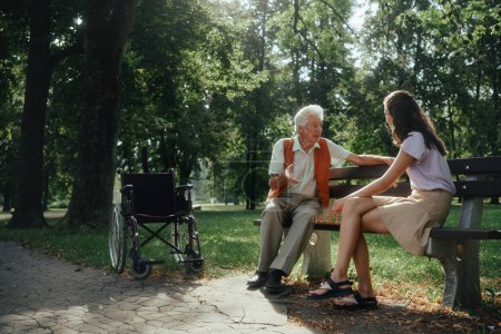 Téléchargez les photos : Une jeune aidante jouant aux échecs avec un aîné solitaire dans le parc de la ville. Le vieil homme en fauteuil roulant passe du temps à l'extérieur avec sa petite-fille, jouant aux échecs et bavardant. - en image libre de droit