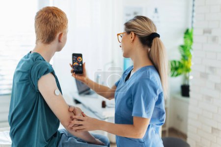 Photo for Diabetologist doctor teaching young patient ho to connect continuous glucose monitor sensor to his smartphone. Doctor checking teenage boys glucose data in the real time. Endocrinologist taking care - Royalty Free Image