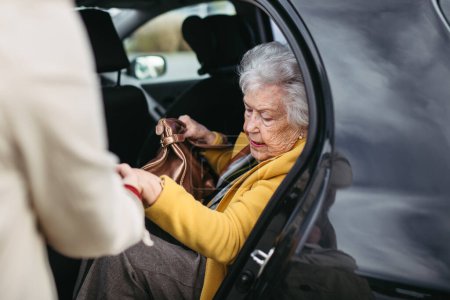 Téléchargez les photos : Vieille dame sortant de la voiture, soignante l'aidant, lui tenant la main. Femme âgée a du mal à se lever du siège arrière de la voiture. - en image libre de droit