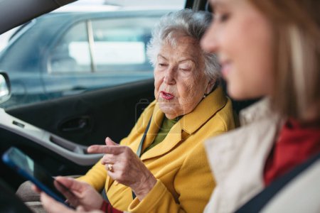 Foto de Nieta conduciendo a su abuela anciana en el coche, llevándola al médico, de compras o al banco. Cuidador de conducir anciana a la iglesia, farmacia, en busca de una ruta en un teléfono inteligente. - Imagen libre de derechos