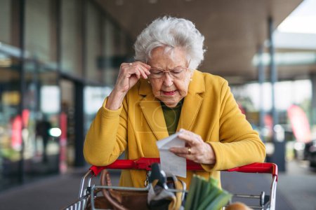 Téléchargez les photos : Une femme âgée vérifiant son reçu après un achat, regardant le montant de l'argent dépensé, s'assurant que toutes les charges sont correctes. - en image libre de droit
