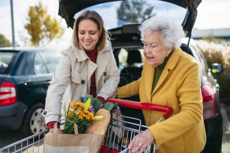Foto de Nieta madura ayudando a su abuela a cargar los comestibles en el coche. Mujer mayor de compras en el centro comercial. - Imagen libre de derechos