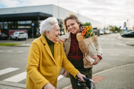 Photo for Mature granddaughter carrying grandmothers shopping bag. Senior woman and caregiver going to home with goceries from the supermarket, during cold autumn day. - Royalty Free Image