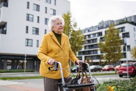 Foto de Una mujer mayor con un caminante de movilidad caminando por las calles de la ciudad durante el día de otoño, disfrutando del hermoso clima soleado. Anciana saboreando cada momento, viviendo la vida al máximo. - Imagen libre de derechos