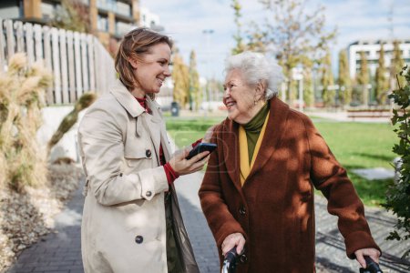 Photo for Front view of grandmother and mature granddaughter on a walk in the city park, during a windy autumn day. Caregiver and senior lady with mobility walker talking and enjoying the fall weather. - Royalty Free Image