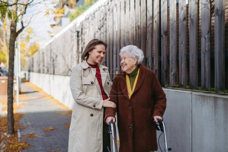 Photo for Front view of grandmother and mature granddaughter on a walk in the city park, during a windy autumn day. Caregiver and senior lady with mobility walker enjoying the fall weather. - Royalty Free Image