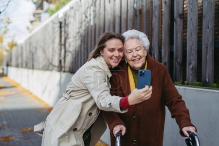 Téléchargez les photos : Grand-mère et petite-fille mature en promenade dans les rues de la ville, en prenant selfie. Aide familiale et dame âgée profitant du temps venteux d'automne. - en image libre de droit