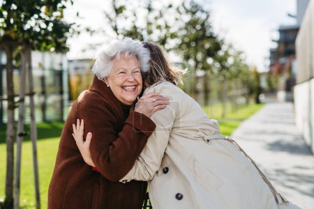 Téléchargez les photos : Grand-mère et petite-fille mûre lors d'une promenade dans le parc de la ville, lors d'une journée d'automne venteuse. Aide familiale et dame âgée profitant du temps de l'automne, se serrant dans leurs bras. - en image libre de droit