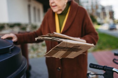 Téléchargez les photos : Femme âgée jetant du carton dans la poubelle du conteneur à déchets devant son complexe d'appartements. - en image libre de droit