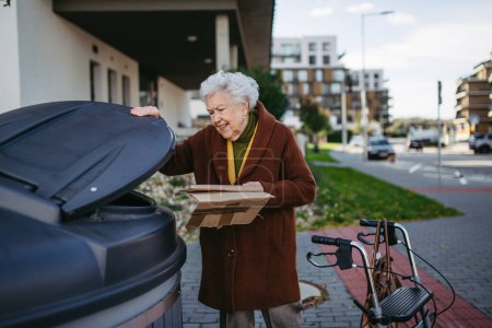 Photo for Elderly woman disposing cardboard into the garbage can of waste container in front of her apartment complex. - Royalty Free Image