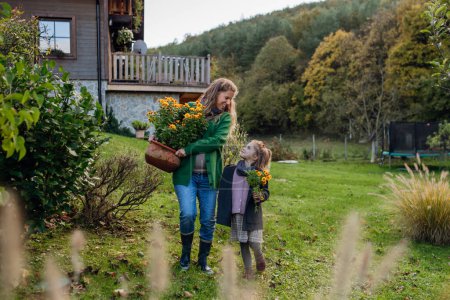 Téléchargez les photos : Mère et fille s'occupent du jardin de la maison, replantent des fleurs. La mère enceinte passe du temps à l'extérieur pendant une froide journée d'automne. - en image libre de droit