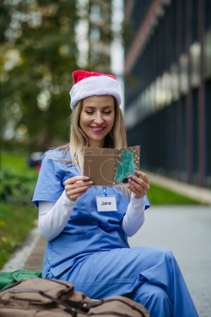 Foto de Enfermera con sombrero de navidad en la cabeza leyendo la tarjeta de navidad del paciente infantil. Trabajando en el hospital el día de Navidad. Doctora trabajando un turno de Navidad y no puede estar con su familia. - Imagen libre de derechos