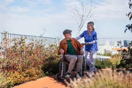 Foto de Enfermera empujando a un hombre mayor en silla de ruedas. Mujer cuidadora y anciano disfrutando de un cálido día de otoño en un hogar de ancianos, hogar de cuidado residencial. - Imagen libre de derechos