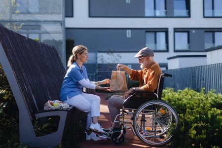 Foto de Enfermera almorzando o cenando al aire libre con un hombre mayor en silla de ruedas. Mujer cuidadora y anciano disfrutando de un cálido día de otoño en un hogar de ancianos, haciendo picnic. - Imagen libre de derechos