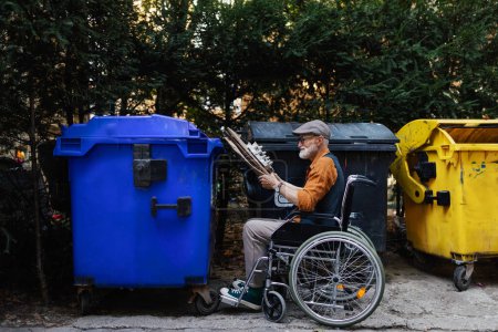 Photo for Senior man in wheelchair throwing paper waste, cardboard into recycling container in front his apartment complex. Elderly man sorting the waste according to material into colored bins. - Royalty Free Image