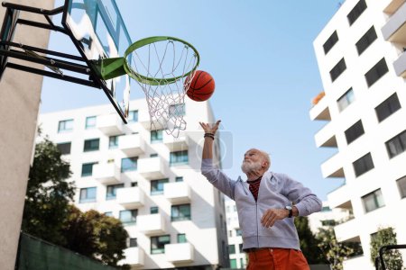 Photo for Senior man playing basketball outdoors on basketball court in the city, in community center. An older, vital man has active lifestyle, doing sport every day. - Royalty Free Image