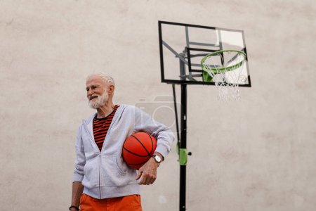 Téléchargez les photos : Homme âgé jouant au basket-ball en plein air sur le terrain de basket dans la ville, dans le centre communautaire. Un homme plus âgé et vital a un mode de vie actif, faisant du sport tous les jours. - en image libre de droit