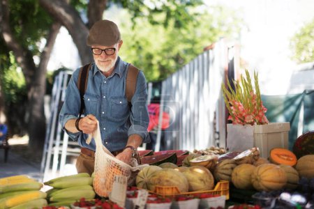 Téléchargez les photos : Portrait d'un homme âgé faisant du shopping au marché de la ville. Homme âgé achetant des légumes et des fruits frais au stand du marché. - en image libre de droit