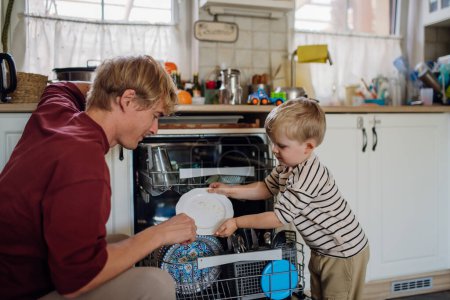 Téléchargez les photos : Petit garçon aidant père à charger le lave-vaisselle après le petit déjeuner ensemble. Nettoyage de la cuisine avant de partir au travail et garderie. routine matinale familiale. - en image libre de droit