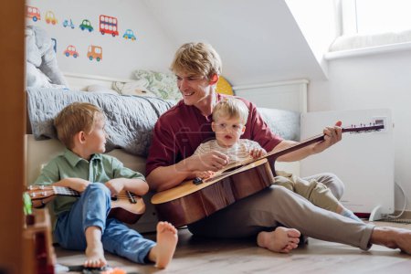 Photo for Father teaching boys to play on guitar. Boys having fun in their room with dad, playing guitar and singing. Concept of Fathers Day, and fatherly love. - Royalty Free Image