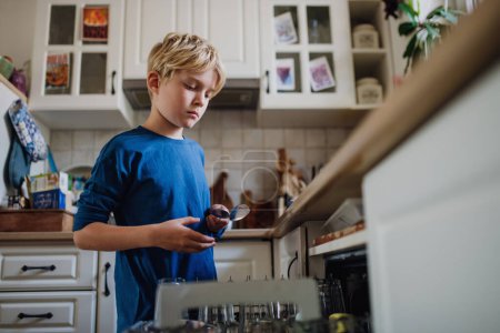Téléchargez les photos : Jeune garçon aidant à charger le lave-vaisselle après le petit déjeuner. Nettoyage de la cuisine avant de partir à l'école. routine matinale familiale. - en image libre de droit
