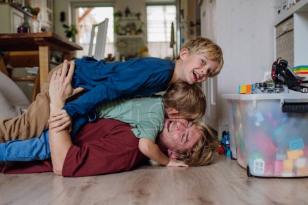 Téléchargez les photos : Portrait de père embrassant ses deux fils à la maison, s'amusant, couché sur le sol et riant. Papa et les garçons passent du bon temps ensemble, coller. - en image libre de droit