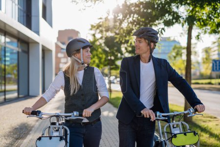 Photo for Colleagues commuting through the city, talking and walking by bike on street. Middle-aged city commuters traveling from work by bike after a long workday. Husband and wife riding bikes in city. - Royalty Free Image