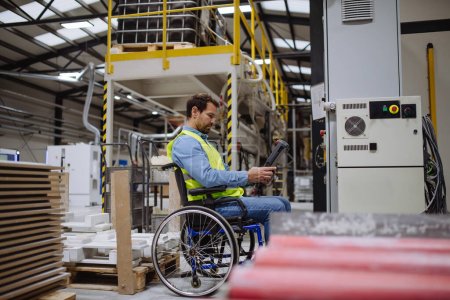 Téléchargez les photos : Homme en fauteuil roulant travaillant dans une usine industrielle moderne, dans un poste de travail réglable. Concept de travailleurs handicapés, lieu de travail accessible pour les employés à mobilité réduite. - en image libre de droit