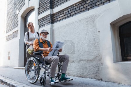 Photo for Granddaughter pushing senior man in wheelchair on street. Buying newspaper in newsstand. Female caregiver and elderly man enjoying a warm autumn day, going home from shopping trip. - Royalty Free Image