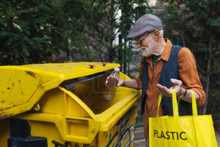 Téléchargez les photos : Homme âgé jetant des déchets plastiques, bouteilles dans un conteneur de recyclage en face de son appartement. Homme âgé triant les déchets selon le matériau dans des poubelles colorées. - en image libre de droit