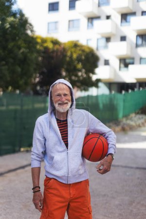 Foto de Hombre mayor jugando al baloncesto al aire libre en la cancha de baloncesto en la ciudad, en el centro comunitario. Un hombre mayor y vital tiene un estilo de vida activo, haciendo deporte todos los días.. - Imagen libre de derechos