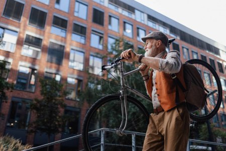 Téléchargez les photos : Un homme âgé, cycliste voyageant à travers la ville en vélo, portant le vélo sur l'épaule. Aîné banlieue en vélo. - en image libre de droit