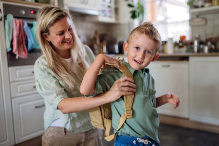 Téléchargez les photos : Mère dit au revoir à son fils avant l'école, l'aidant à mettre son sac à dos sur son dos dans la cuisine. - en image libre de droit