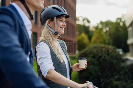 Photo for Beautiful middle-aged woman commuting through the city, buying, drinking coffe in front of office. Female city commuter with helmet traveling from work by bike after a long workday. - Royalty Free Image