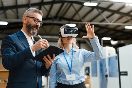 Téléchargez les photos : Les ingénieurs parlent projet en utilisant VR, dans l'usine industrielle moderne. Réalité virtuelle et technologie dans l'industrie manufacturière, lunettes intelligentes guidant le gestionnaire féminin et masculin dans le processus et le rabotage. - en image libre de droit