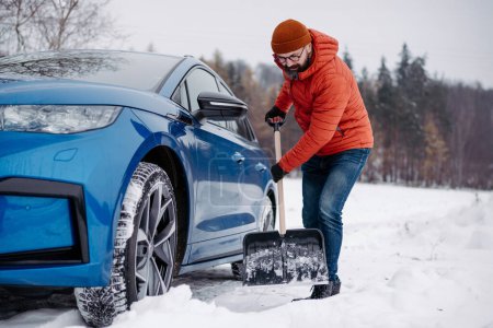 Téléchargez les photos : Un homme libérant sa voiture coincée dans la neige avec une pelle, sortant la voiture de la dérive de neige. Ramasser et jeter de la neige, pelleter. - en image libre de droit