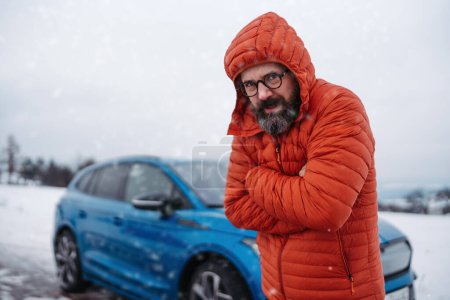 Photo for Angry man standing by electric car, battery run out of power before reaching destination, during snowy winter day. Frustrated man is cold, waiting for help, breakdown service car, tow truck. - Royalty Free Image